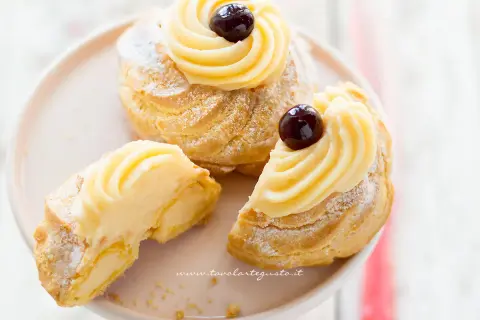 Zeppole di San Giuseppe, dolci soffici con crema pasticcera e ciliegina, uno è tagliato per mostrarne l'interno goloso.