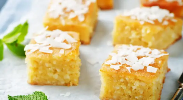 Fette quadrate di torta al cocco, guarnite con scaglie di cocco e foglie di menta fresca.