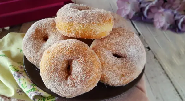 Graffe senza patate, soffici e dorate, ricoperte di zucchero. Un dolce invitante su un piatto scuro, con fiori viola sullo sfondo.