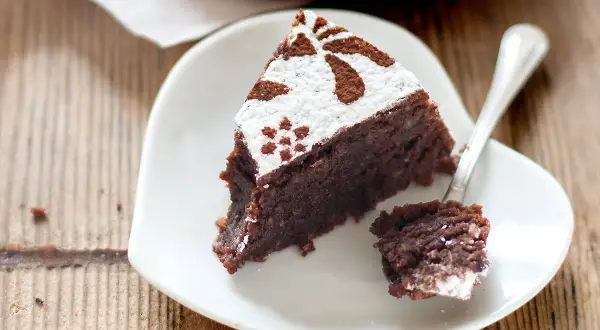 Una fetta di torta caprese ricetta facile, cosparsa di zucchero a velo e cacao, su un piatto bianco a forma di cuore. Un cucchiaino ha prelevato un pezzetto.