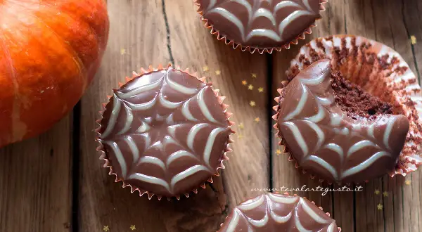 Muffin Halloween facili con decorazione a ragnatela. Una zucca arancione e stelline dorate completano la scena.
