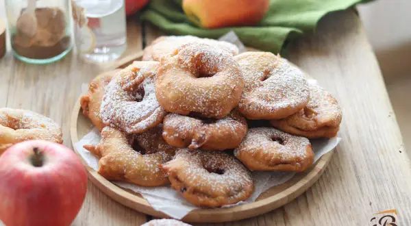 Frittelle di mele della nonna, dorate e cosparse di zucchero a velo, su un piatto di legno, con mele fresche e spezie sullo sfondo.