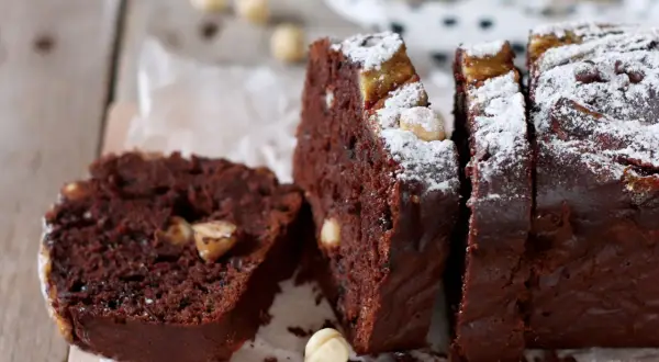 Fetta di banana bread cacao con nocciole e zucchero a velo, su tagliere di legno.