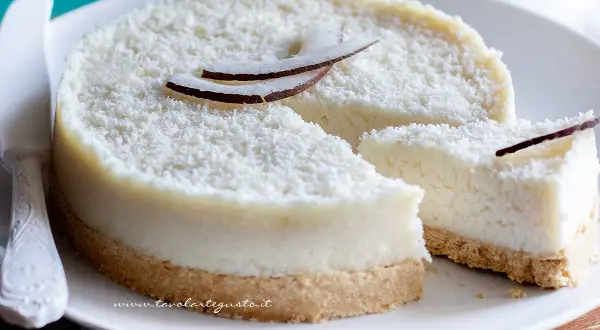 Delizioso cheesecake al cocco, ricoperto di cocco grattugiato e decorato con scaglie di cocco fresco. Una fetta è già stata tagliata.