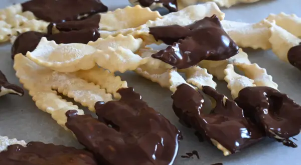 Chiacchiere al cioccolato, dolci croccanti e friabili, cosparse di zucchero a velo e immerse in golosa cioccolata fondente.