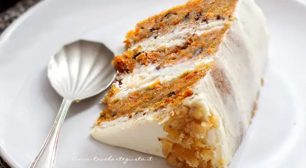 Una fetta di torta di carote, soffice e profumata, con glassa bianca e noci tritate. Accanto, un cucchiaino d'argento.