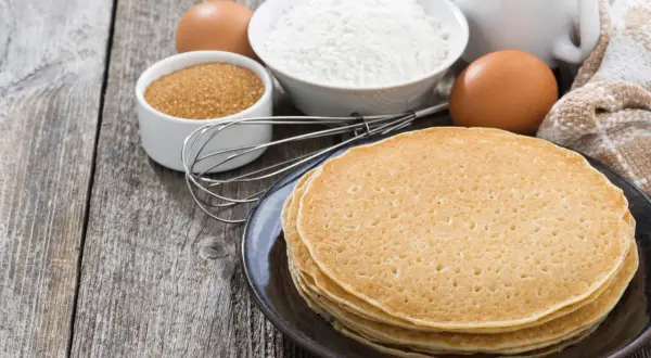 Ingredienti per la ricetta crepes: farina, uova, zucchero, latte e frusta su un tavolo di legno.