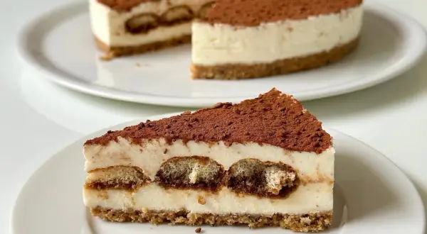 Una fetta di cheesecake tiramisù, con savoiardi inzuppati e cacao in polvere, su un piatto bianco.