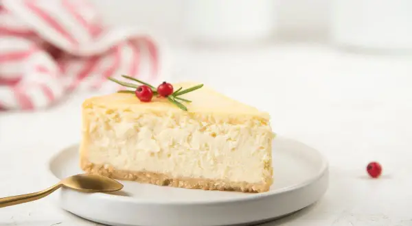 Una fetta di cheesecake con yogurt greco, guarnita con bacche rosse e rosmarino, su un piatto bianco con un cucchiaio dorato.