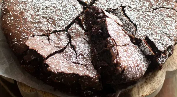 Torta con 2 uova e cioccolato fondente, spolverata di zucchero a velo, con una fetta sollevata che rivela la sua morbidezza.