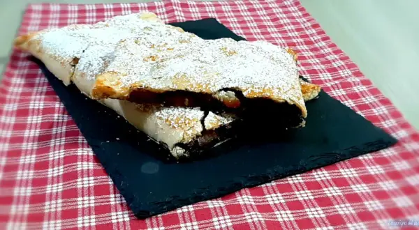 Strudel mele e cioccolato cosparso di zucchero a velo, su una pietra nera e tovaglia a quadri rossi.