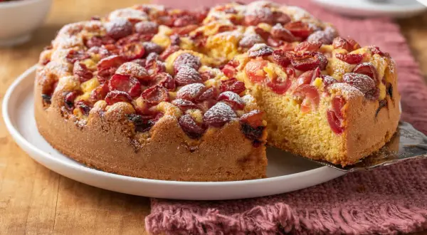 Torta con ciliegie, soffice e profumata, cosparsa di zucchero a velo. Una fetta viene sollevata, mostrando la morbidezza dell'impasto.