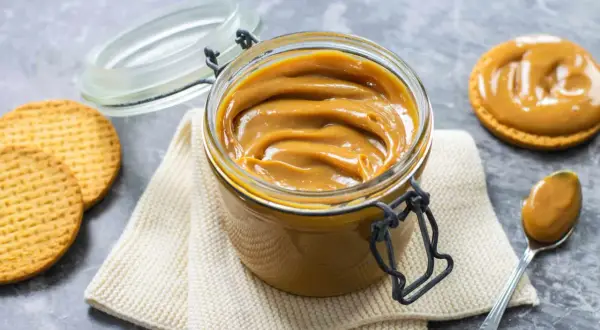 Vaso di vetro pieno di cremoso dulce de leche, con biscotti e un cucchiaino pronto per assaporare questa delizia.