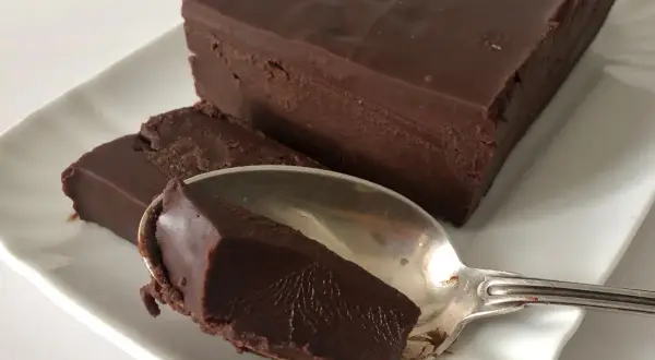 Una mattonella di cioccolato fondente, densa e lucida, su un piatto bianco. Un cucchiaino ne ha prelevato un pezzo.