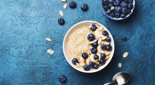 Porridge ricetta: una ciotola di avena con mirtilli, banane e mandorle a lamelle su sfondo blu.
