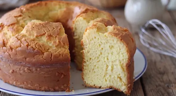 Una torta alta, soffice e dorata, con una fetta tagliata che rivela la sua morbidezza. Sullo sfondo, uova e latte, ingredienti per un dolce fatto in casa.