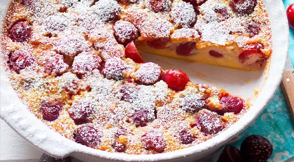 Clafoutis di ciliegie appena sfornato, cosparso di zucchero a velo, con una fetta mancante che rivela la morbida consistenza.