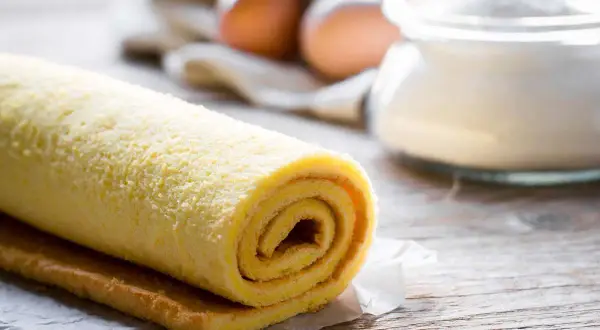 Rotolo di pasta biscotto soffice, pronto per essere farcito. Sullo sfondo, uova e zucchero, ingredienti essenziali per un dolce fatto in casa.