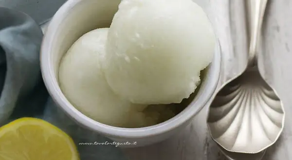 Coppetta con due palline di sorbetto al limone alcolico, accanto una fetta di limone e un cucchiaio decorato.