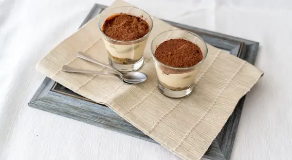 Due bicchierini di tiramisù con colomba, cosparsi di cacao, su un runner di iuta.