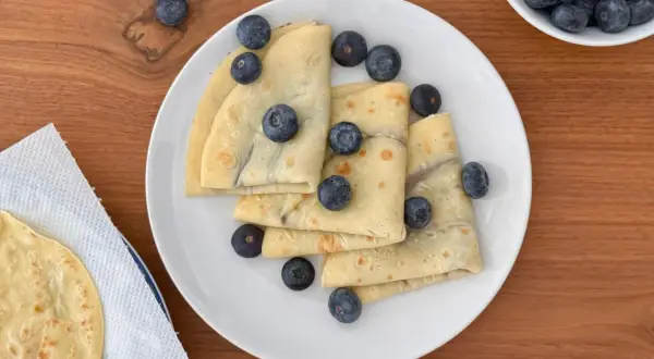 Deliziose crepes all'acqua, piegate e guarnite con mirtilli freschi, su un piatto bianco.