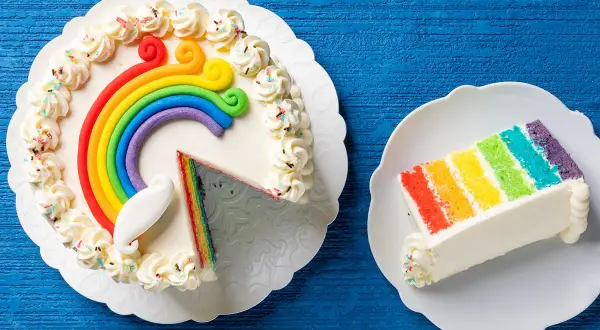 Torta arcobaleno con decorazioni colorate e una fetta servita su un piatto.