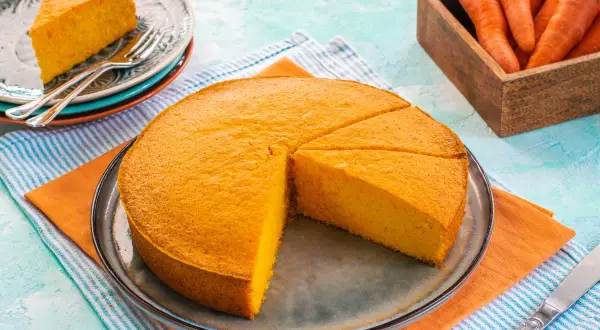 Una soffice torta di carote, appena affettata, su un piatto. Accanto, un piattino con una fetta e delle carote fresche in un cesto.
