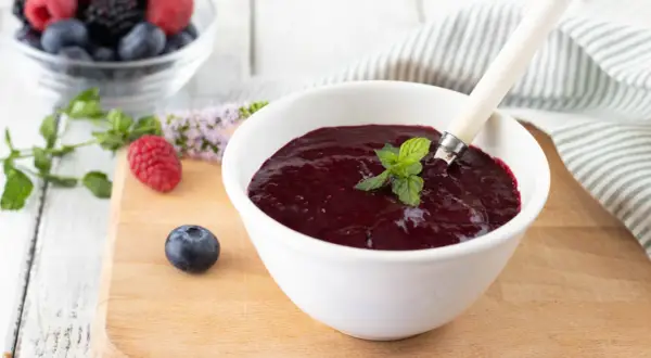 Una ciotola bianca contiene un denso coulis frutti di bosco, guarnito con menta fresca. Frutti di bosco freschi e foglie di menta sono sparsi sul tagliere.