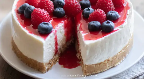 Cheesecake ricetta originale con frutti di bosco freschi e coulis rosso. Una fetta è stata tolta, rivelando la sua consistenza cremosa.