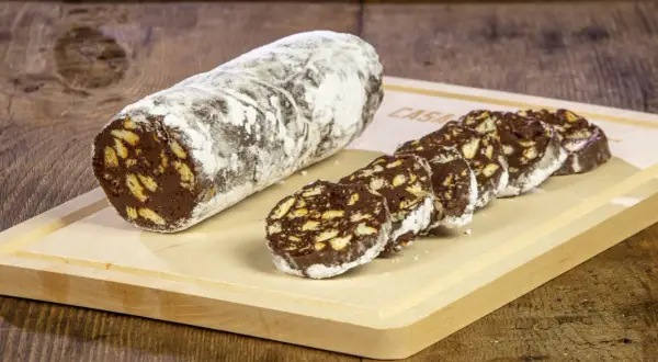 Un rotolo di salame al cioccolato, cosparso di zucchero a velo, è affettato su un tagliere di legno. Pezzi croccanti di biscotto si mescolano al cioccolato fondente.