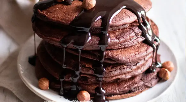 Pancake cacao golosi, impilati e ricoperti da una cascata di salsa al cioccolato fondente e nocciole intere.