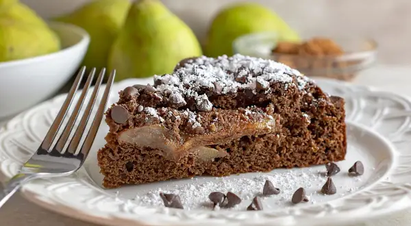 Una fetta di torta pere e gocce di cioccolato, cosparsa di zucchero a velo e gocce di cioccolato, su un piatto bianco con una forchetta.