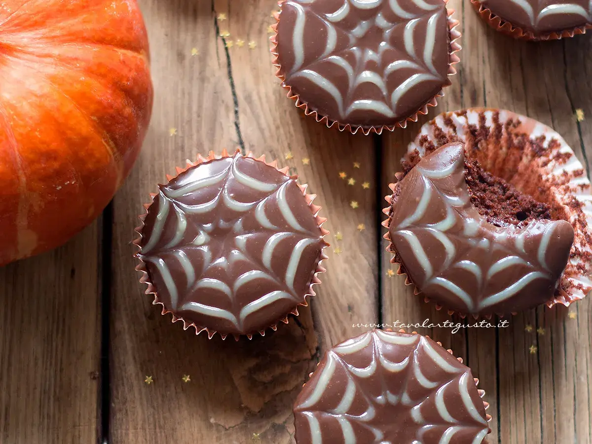 Muffin Halloween facili con decorazione a ragnatela. Una zucca arancione e stelline dorate completano la scena.