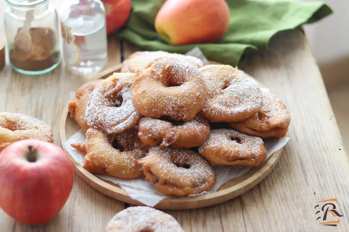 Frittelle di mele della nonna, dorate e cosparse di zucchero a velo, su un piatto di legno, con mele fresche e spezie sullo sfondo.