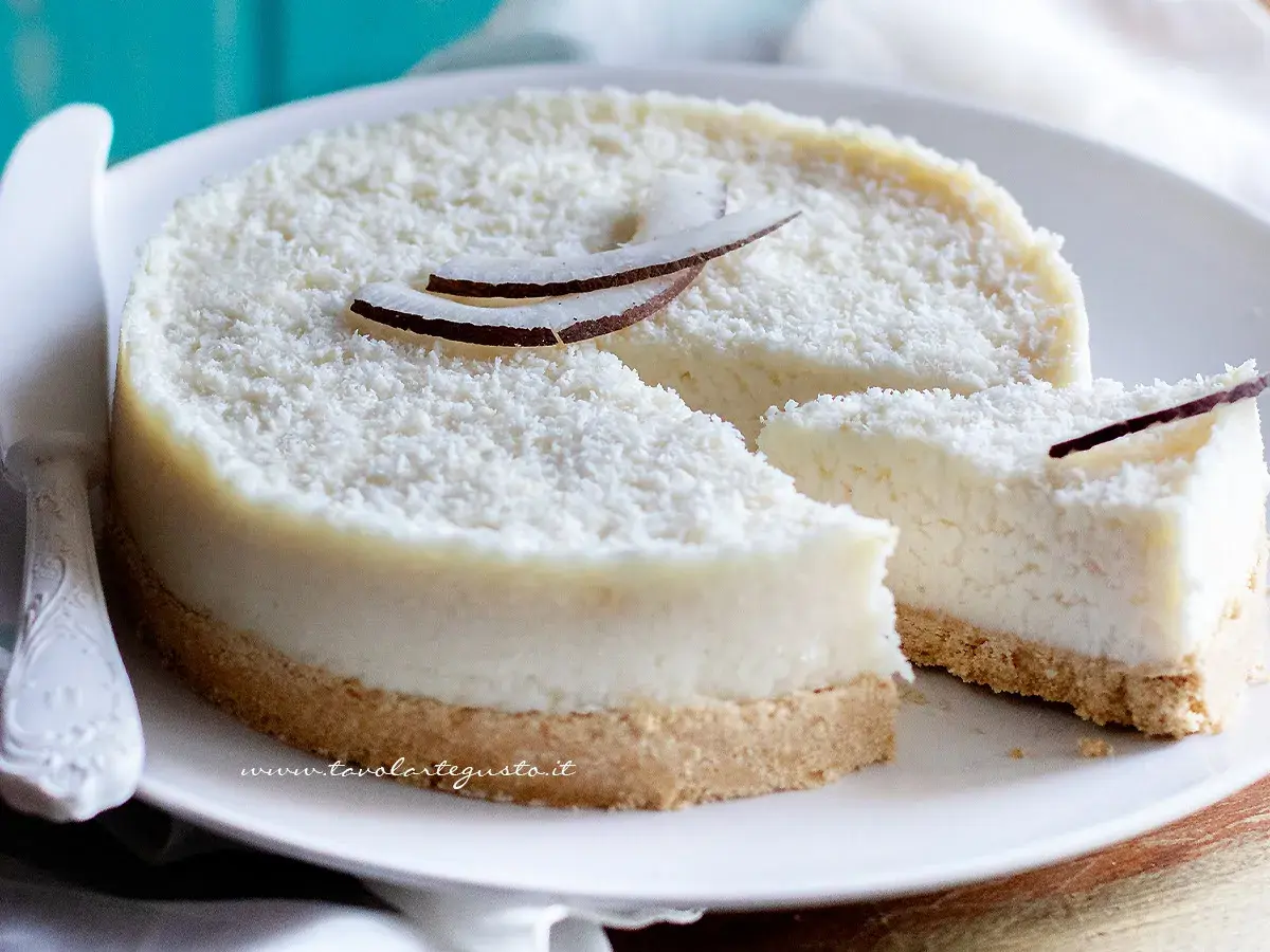 Delizioso cheesecake al cocco, ricoperto di cocco grattugiato e decorato con scaglie di cocco fresco. Una fetta è già stata tagliata.