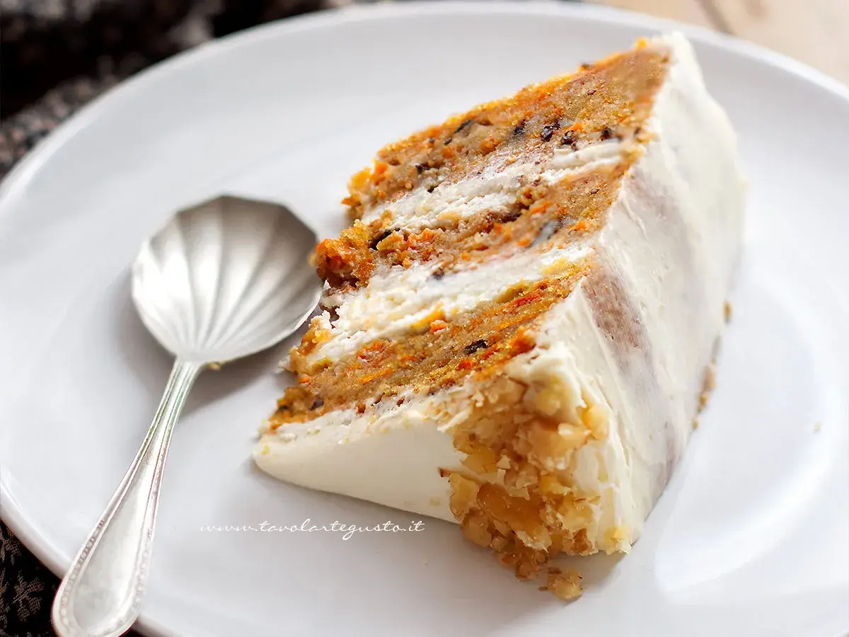 Una fetta di torta di carote, soffice e profumata, con glassa bianca e noci tritate. Accanto, un cucchiaino d'argento.