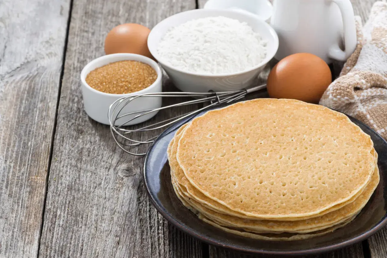 Ingredienti per la ricetta crepes: farina, uova, zucchero, latte e frusta su un tavolo di legno.