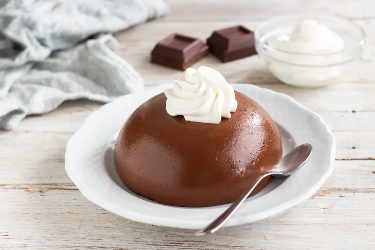 Budino al cacao liscio e lucido, guarnito con panna montata, su un piatto bianco. Accanto, cubetti di cioccolato e una ciotola di panna.