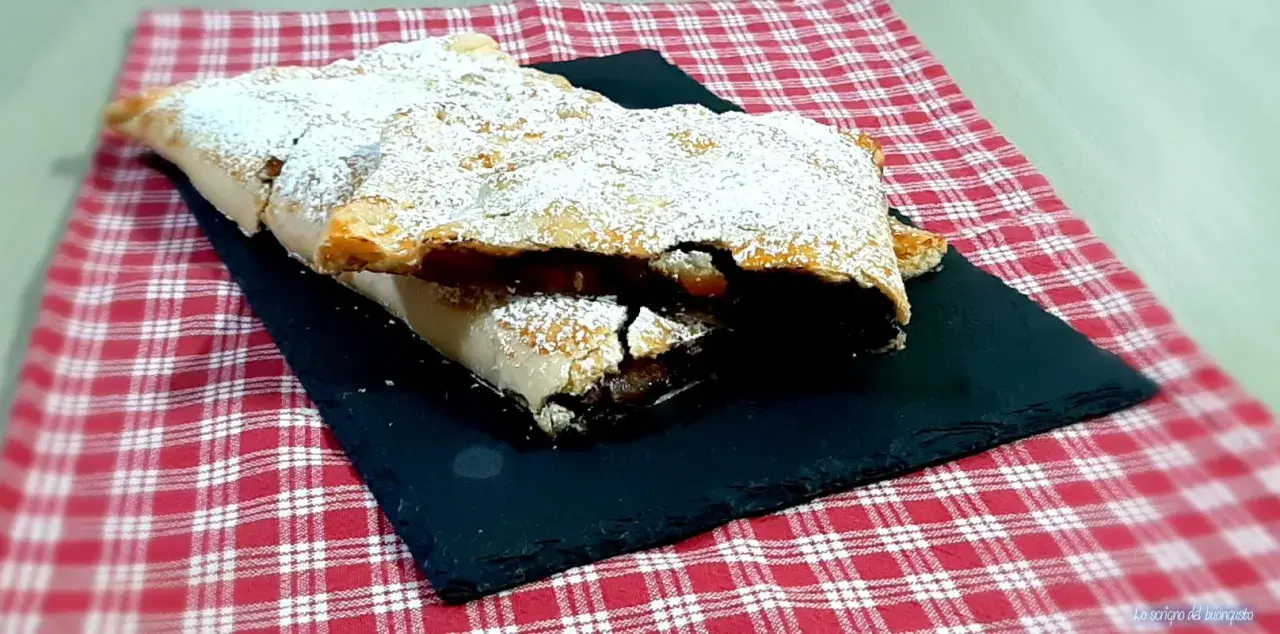 Strudel mele e cioccolato cosparso di zucchero a velo, su una pietra nera e tovaglia a quadri rossi.