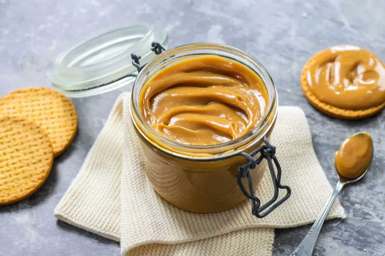Vaso di vetro pieno di cremoso dulce de leche, con biscotti e un cucchiaino pronto per assaporare questa delizia.