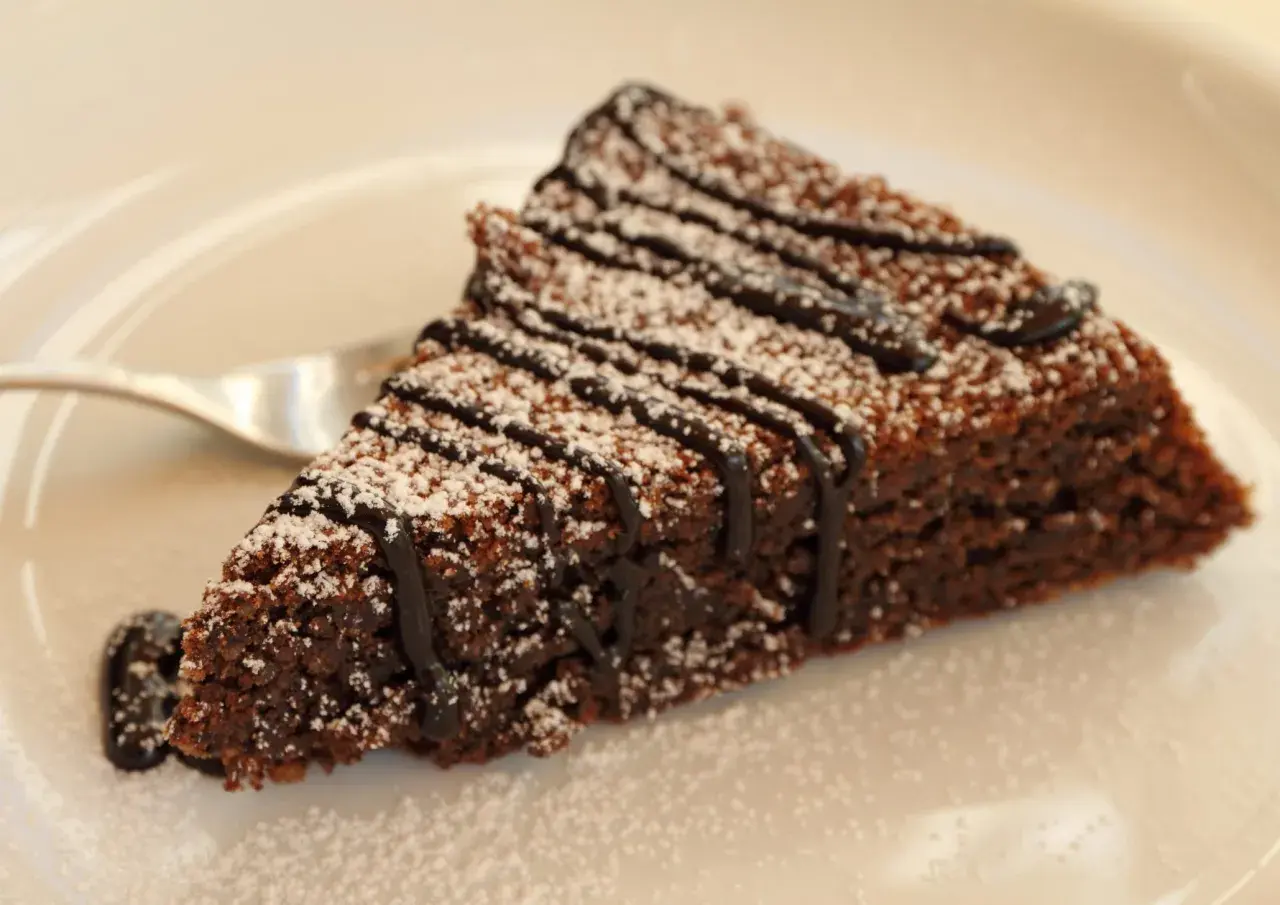 Una fetta di torta caprese al cioccolato, cosparsa di zucchero a velo e con una colatura di cioccolato fondente.