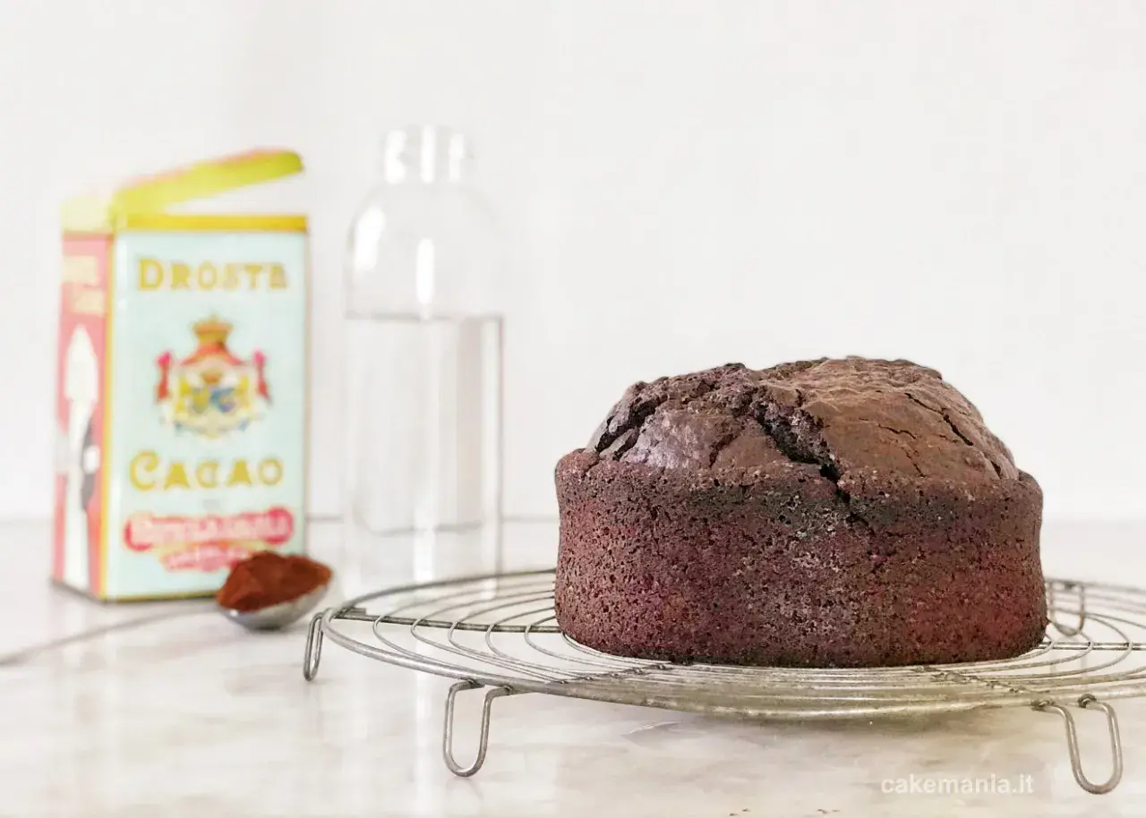 Torta all'acqua al cacao appena sfornata, su una gratella. Accanto, una scatola di cacao Droste e un bicchiere d'acqua.