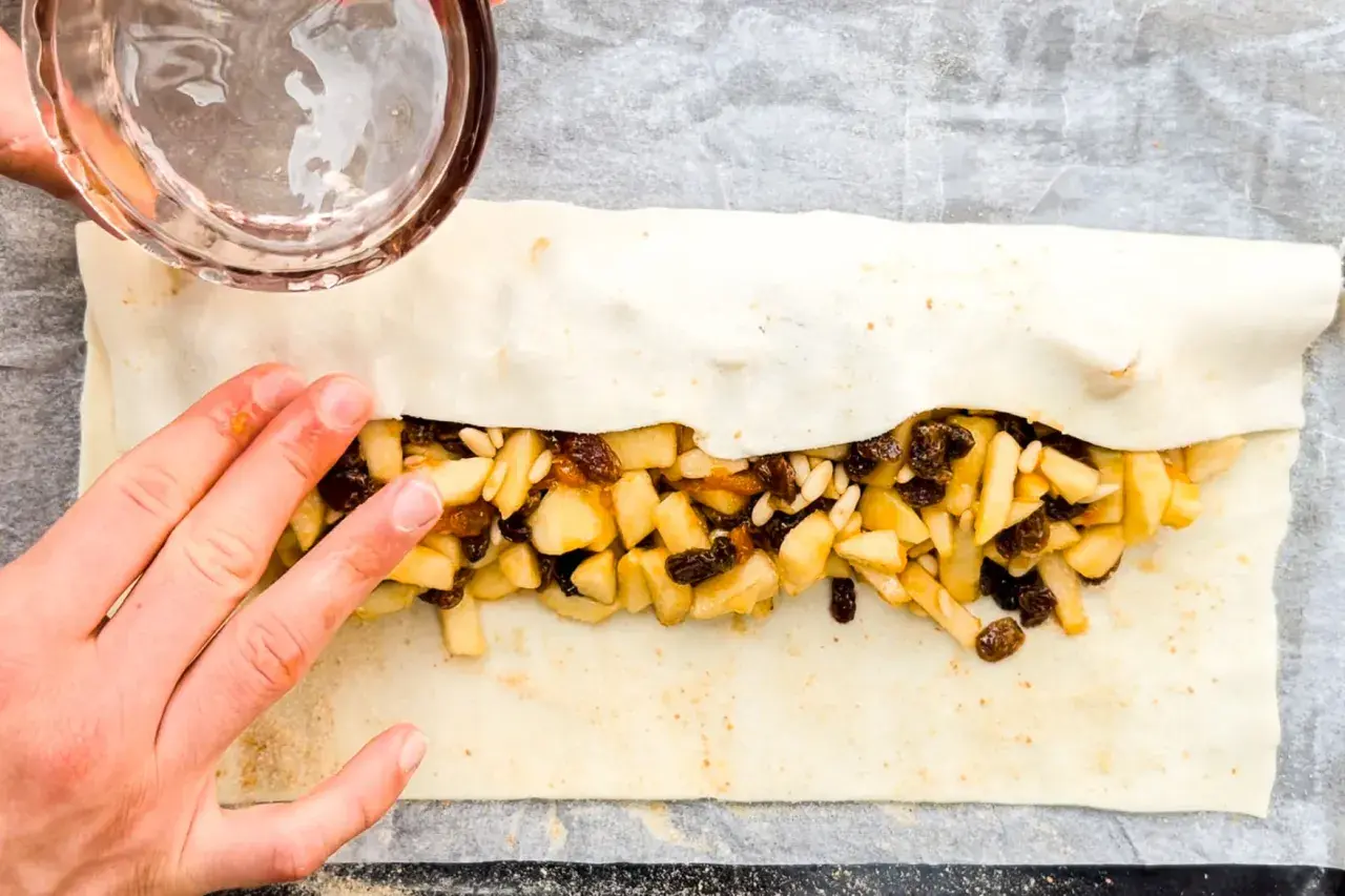 Mani che chiudono uno strudel di mele con pasta sfoglia, ripieno di mele a cubetti, uvetta e pinoli.
