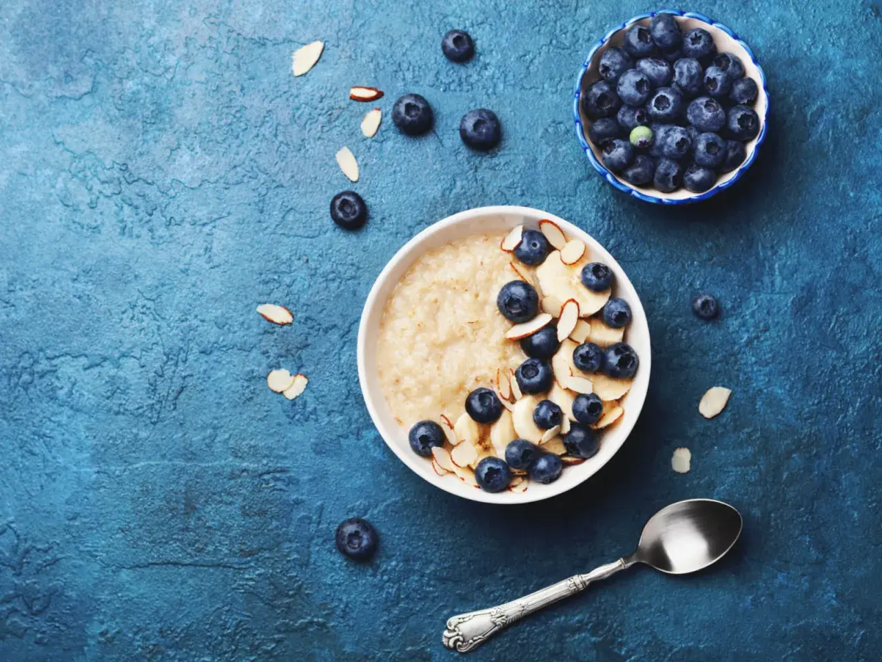 Porridge ricetta: una ciotola di avena con mirtilli, banane e mandorle a lamelle su sfondo blu.