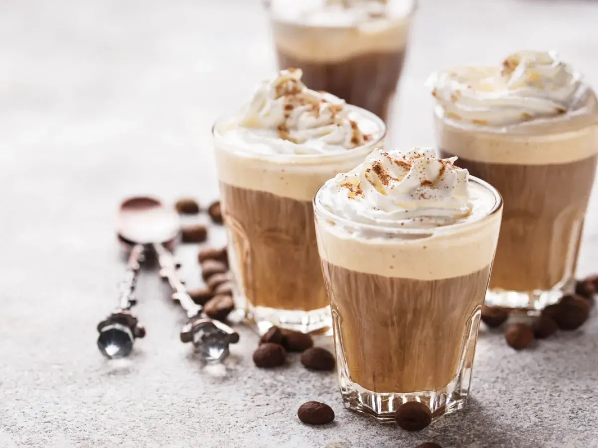 Tre bicchieri di deliziosa crema al caffè, guarniti con panna montata e cacao, circondati da chicchi di caffè.