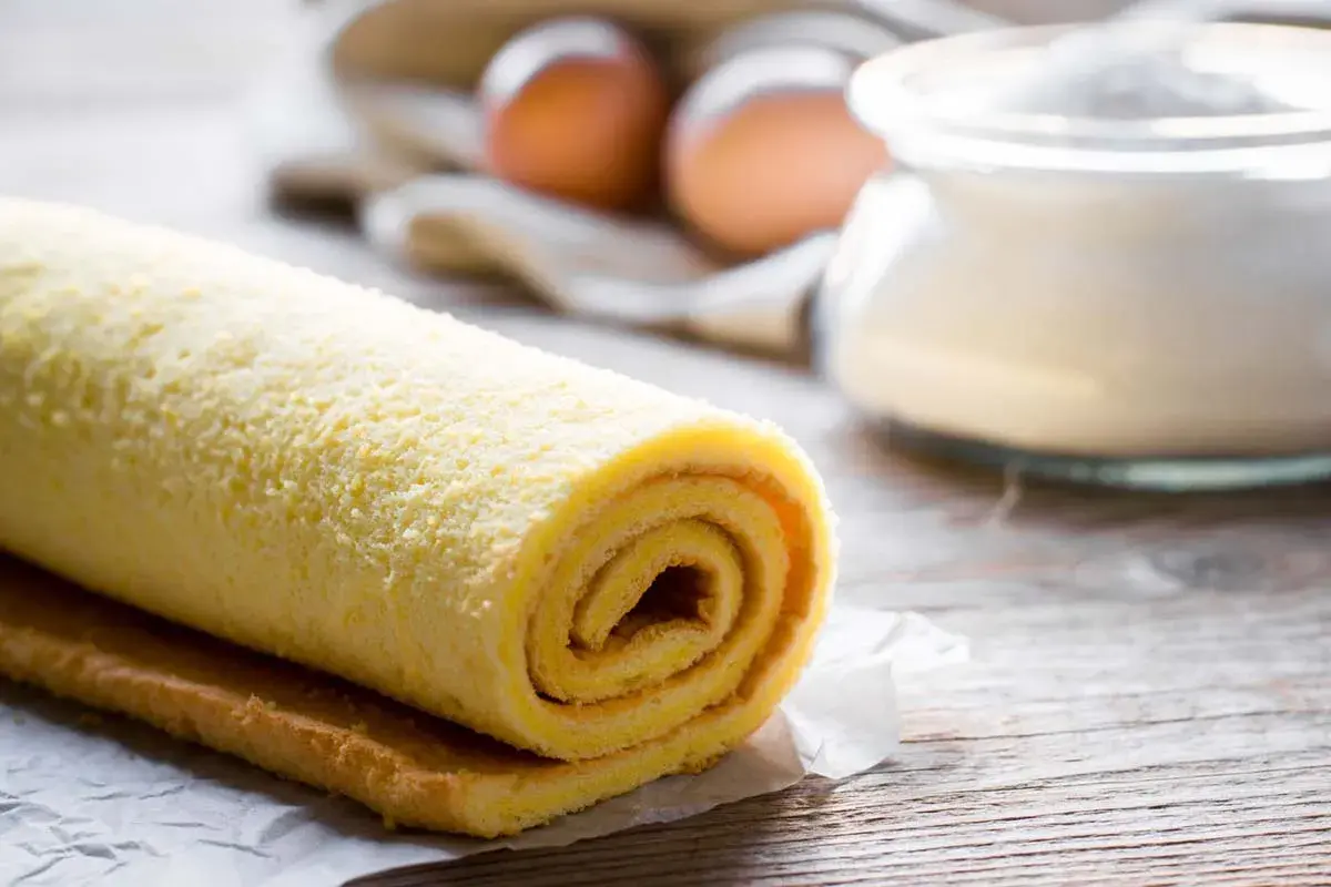 Rotolo di pasta biscotto soffice, pronto per essere farcito. Sullo sfondo, uova e zucchero, ingredienti essenziali per un dolce fatto in casa.