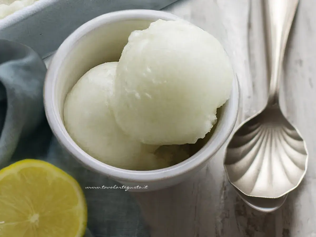 Coppetta con due palline di sorbetto al limone alcolico, accanto una fetta di limone e un cucchiaio decorato.