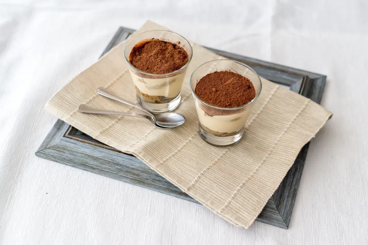 Due bicchierini di tiramisù con colomba, cosparsi di cacao, su un runner di iuta.