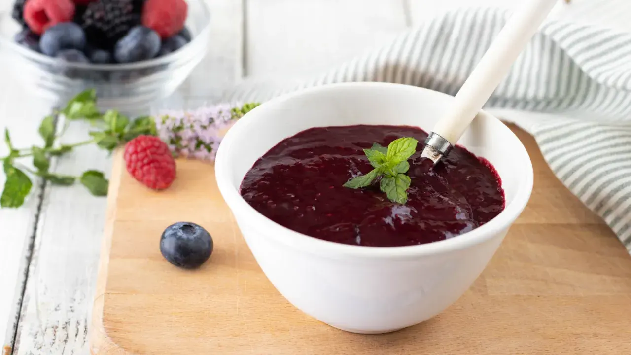 Una ciotola bianca contiene un denso coulis frutti di bosco, guarnito con menta fresca. Frutti di bosco freschi e foglie di menta sono sparsi sul tagliere.