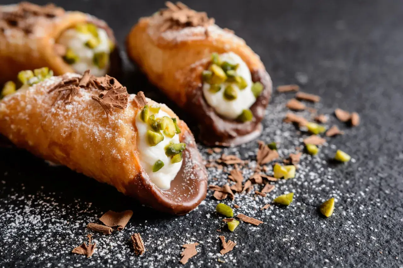 Cannoli siciliani con la loro deliziosa farcitura, guarniti con pistacchi e scaglie di cioccolato su fondo scuro.